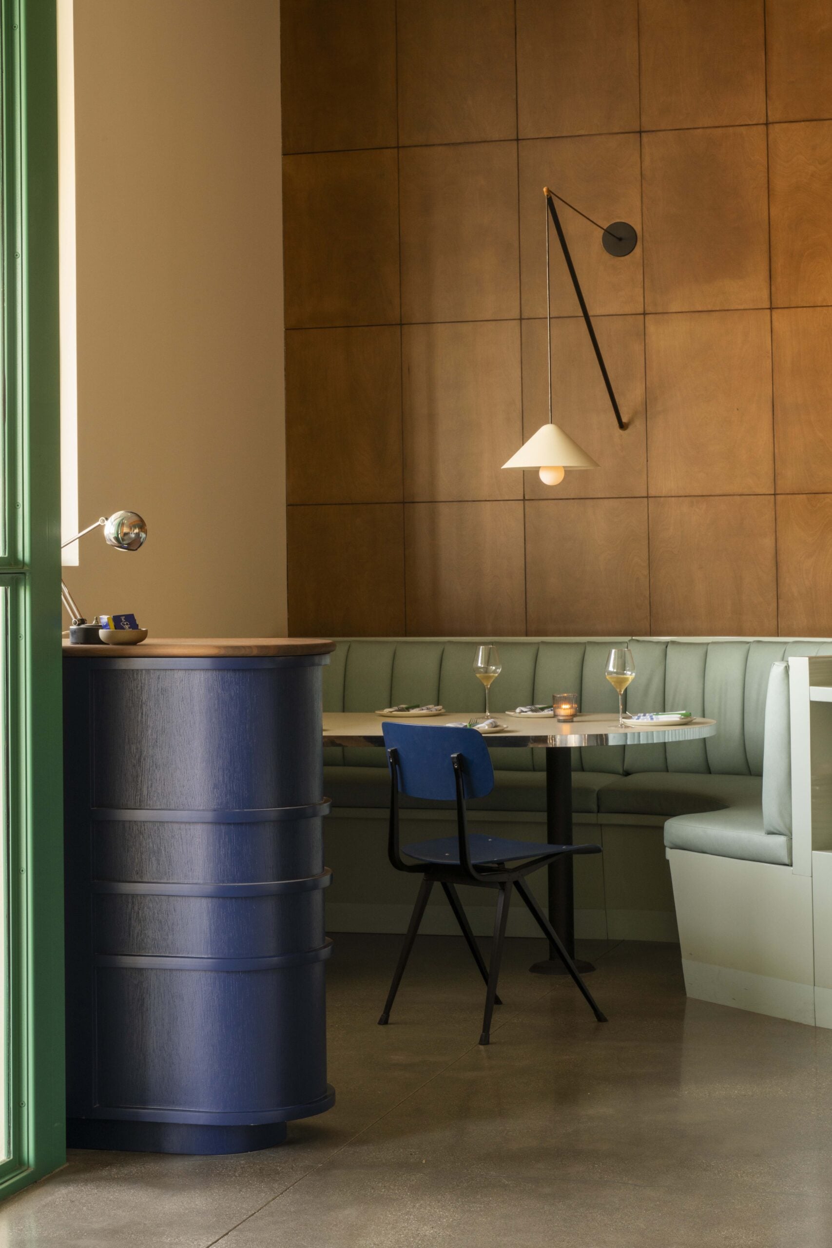 A modern restaurant interior where lovers unite, featuring a wooden wall, geometric pendant light, curved booth with pale green seating, small round table, and a striking blue reception desk.