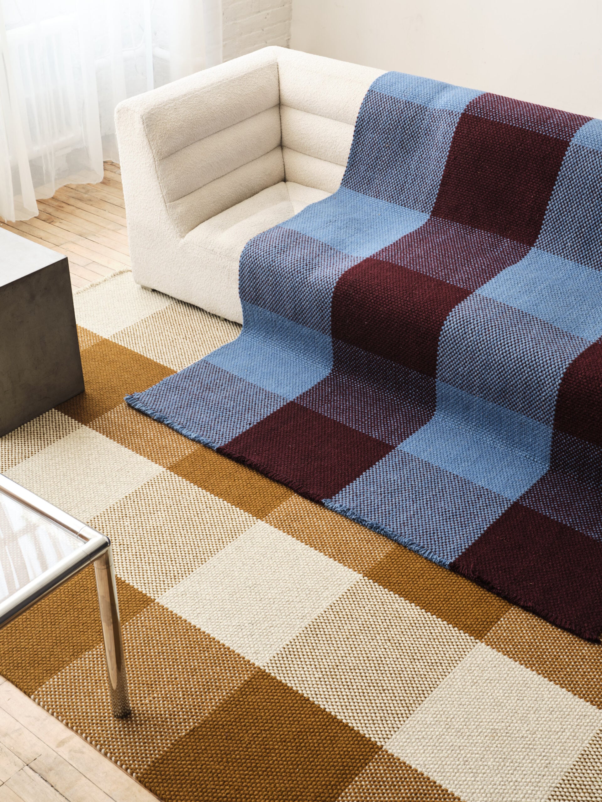 A modern white sofa covered with a large blue and burgundy checkered blanket sits on a tan and cream checkered rug, showcasing stylish textiles in a bright room with wooden floors.