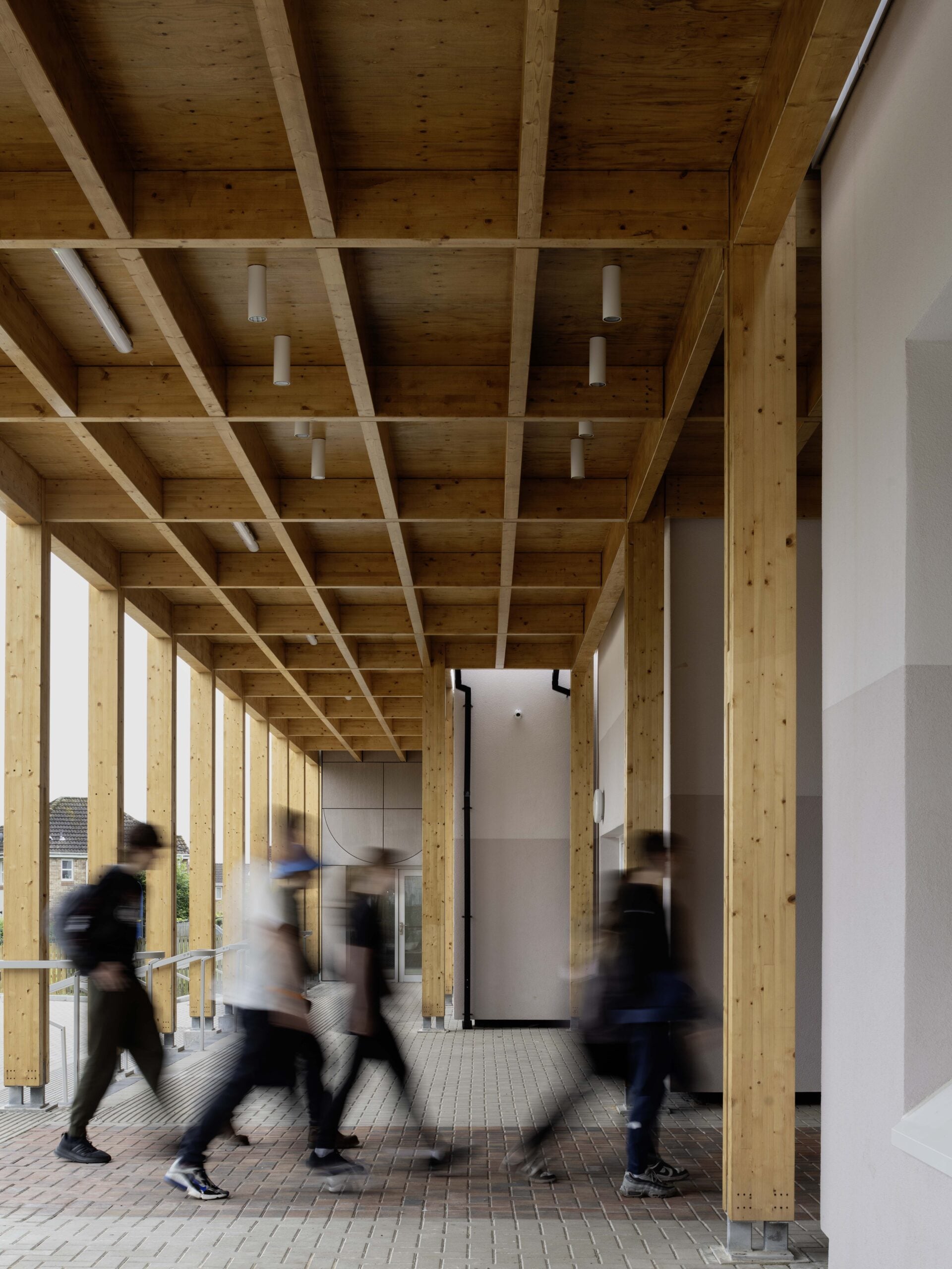A group of people walks under a wooden pergola structure with exposed beams and brick flooring; the image is slightly blurred from their motion.