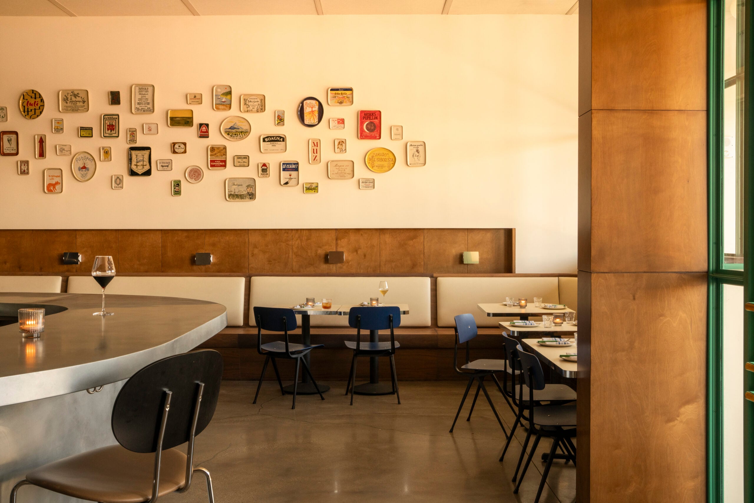 Modern restaurant interior with minimalist decor invites lovers unite over empty tables and chairs, a curved countertop, and a wall display of assorted tins and containers.