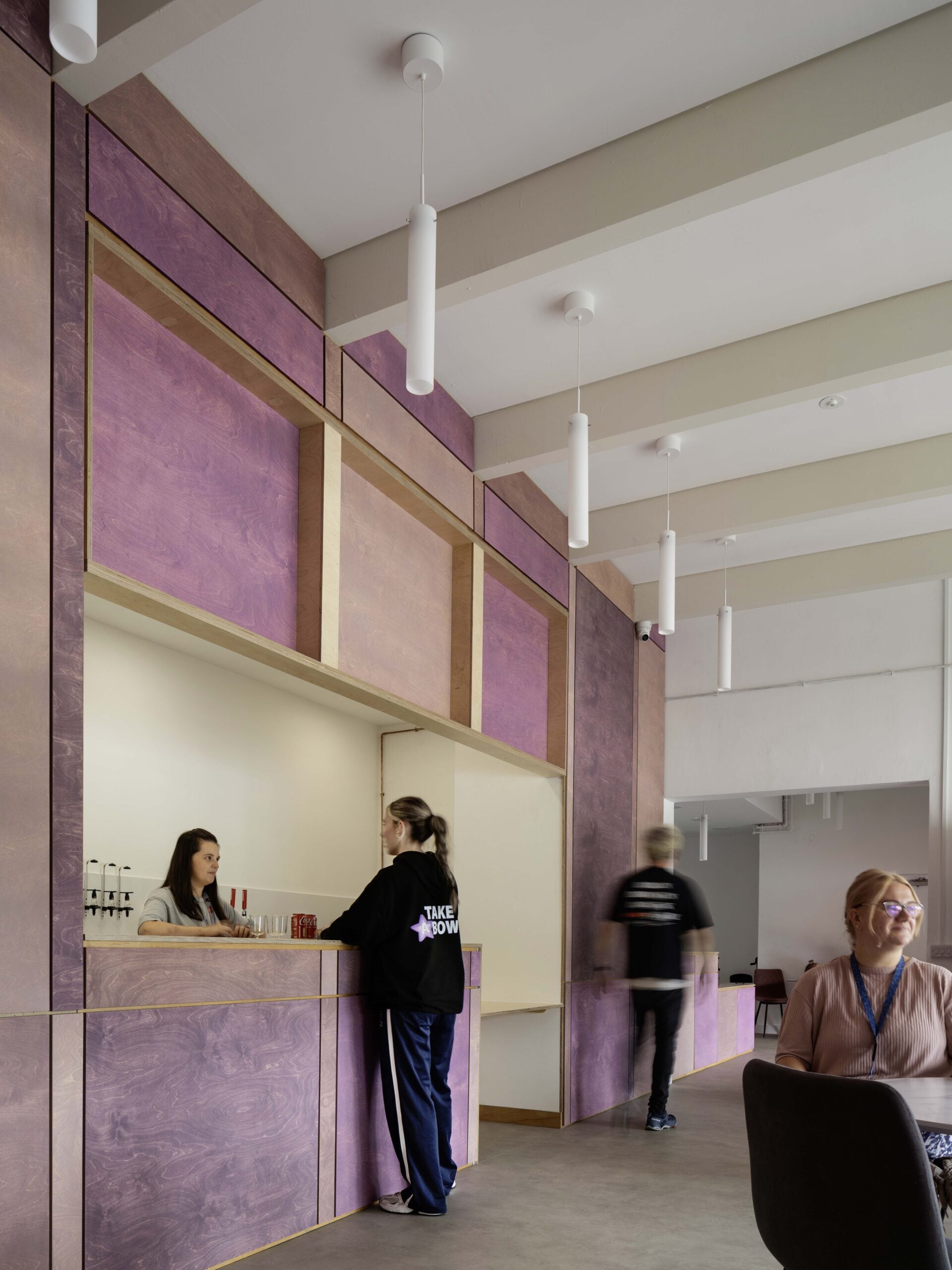 Two people stand at a counter built into a wall with purple wood paneling; a person walks by and another sits at a table in the foreground.