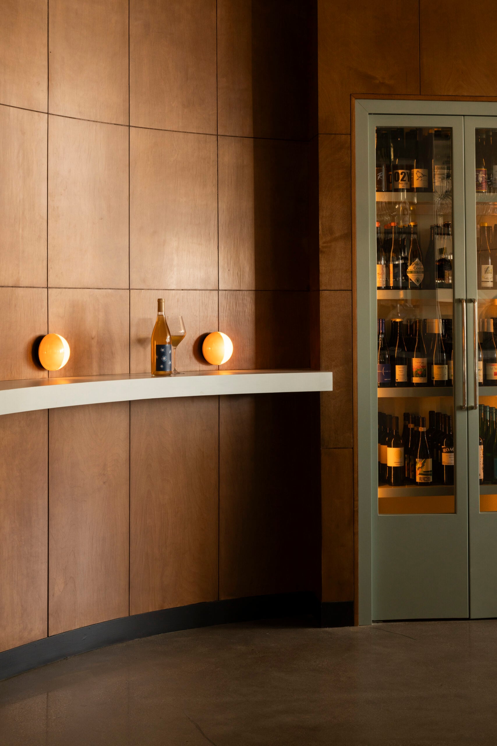 A minimalist bar area with wood-paneled walls, a white shelf holding a wine bottle and two spherical lamps—an inviting space where lovers unite beside a glass cabinet filled with bottles.