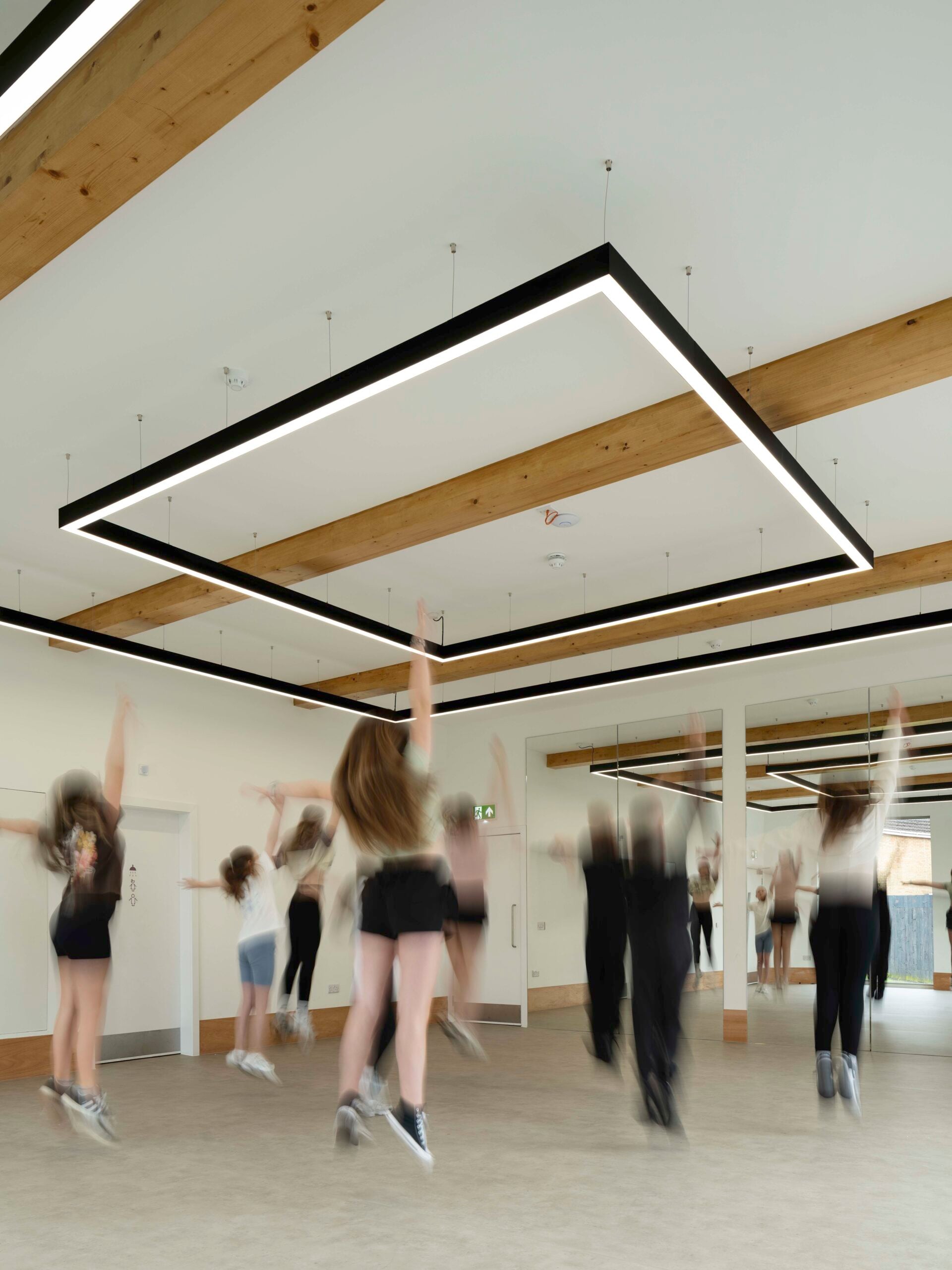 A group of people jumping in a modern, spacious room with large mirrors, wooden beams, and geometric ceiling lights.