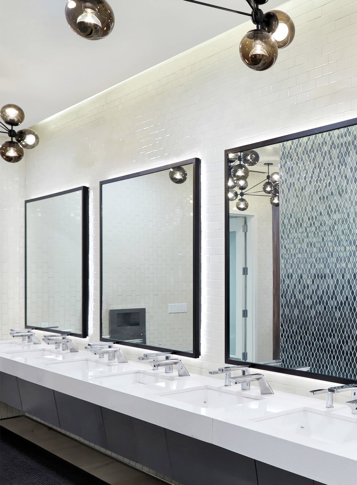 A modern public restroom featuring sleek material finishes, a row of white sinks, large rectangular mirrors, white tiled walls, and contemporary light fixtures.