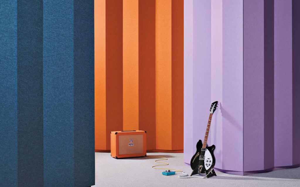 A black electric guitar made from premium material, effects pedal, and orange amplifier are set against large vertical panels in blue, orange, and purple colors.