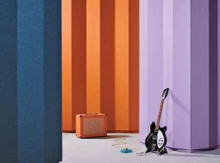 A black electric guitar made from premium material, effects pedal, and orange amplifier are set against large vertical panels in blue, orange, and purple colors.
