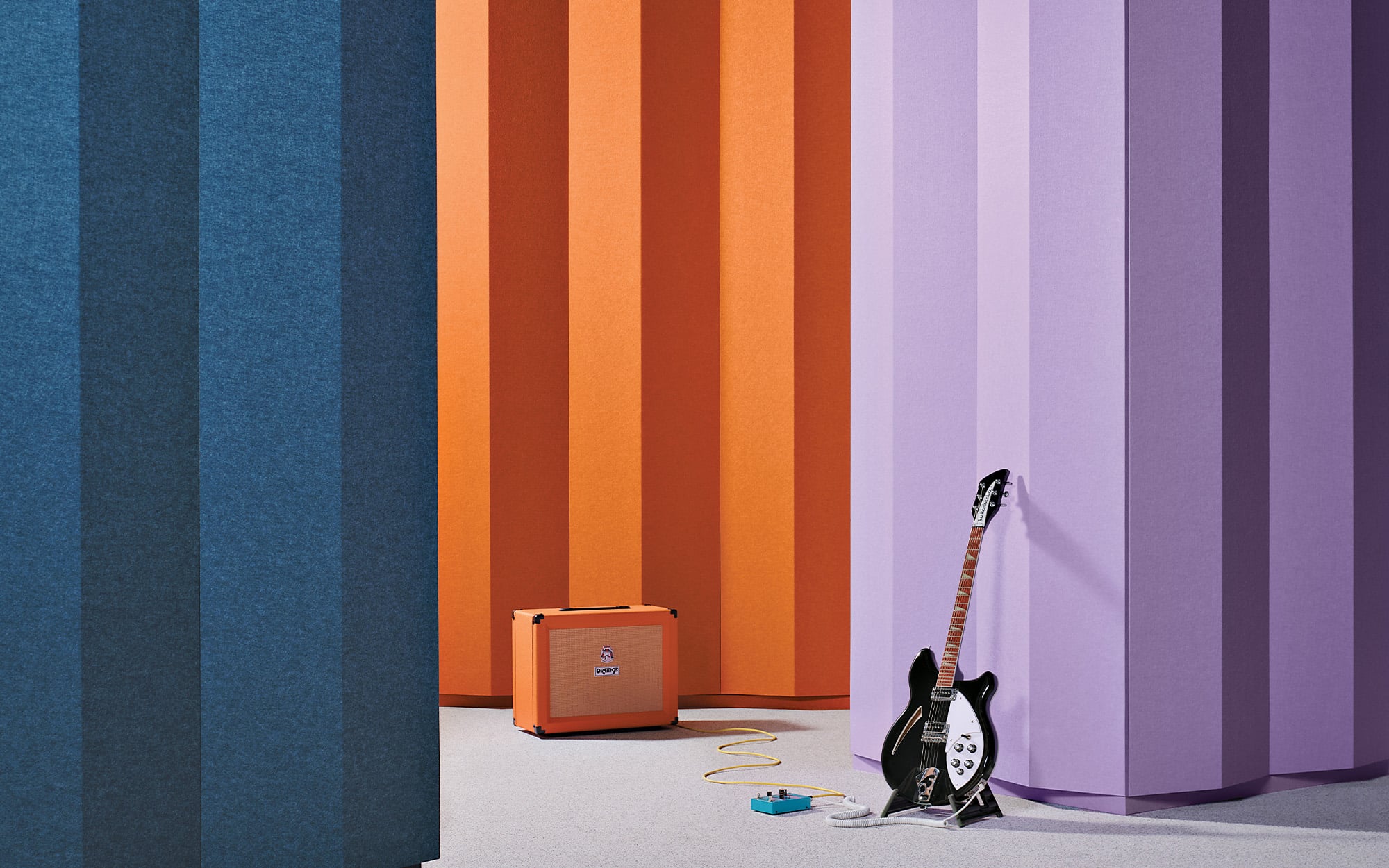 A guitar, effects pedal, and orange amplifier made from quality material are placed on the floor against tall, vertical blue, orange, and purple panels.