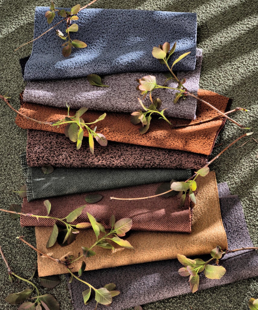 Six folded textured material pieces in different earthy colors are stacked on a green surface, with leafy branches placed on top and around them.