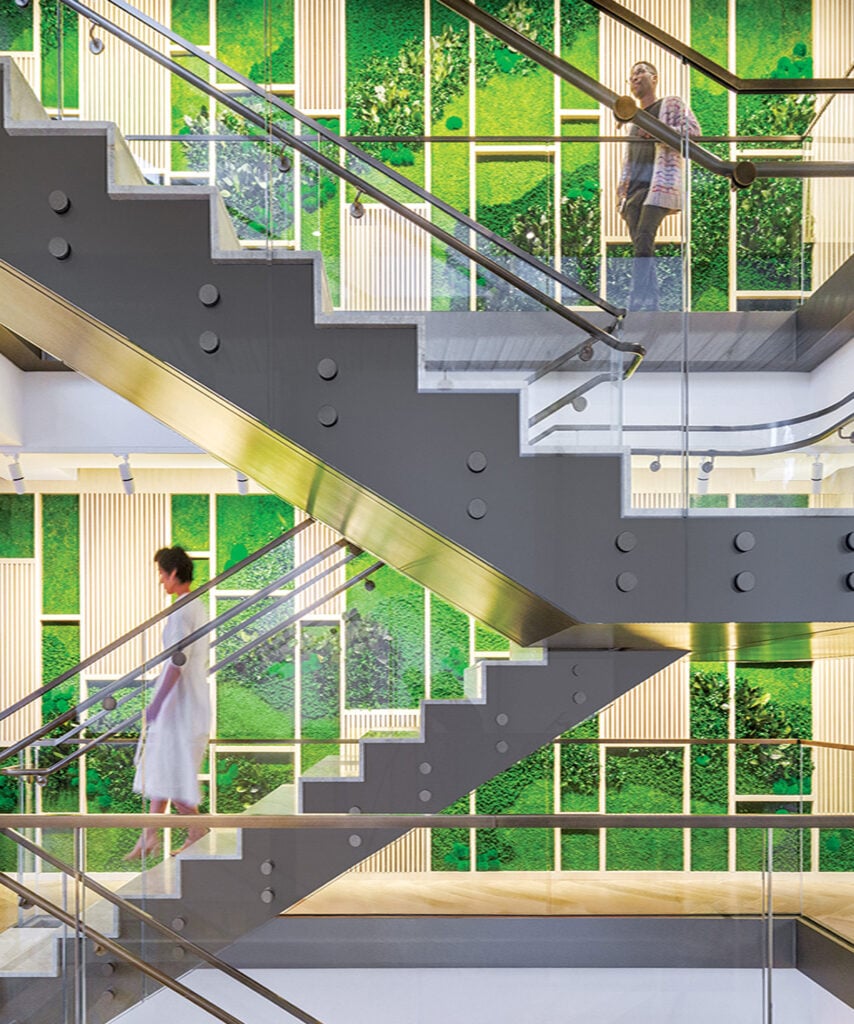 Two people use opposite flights of a modern staircase, showcasing the building’s green and white material wall panels in the bright background.