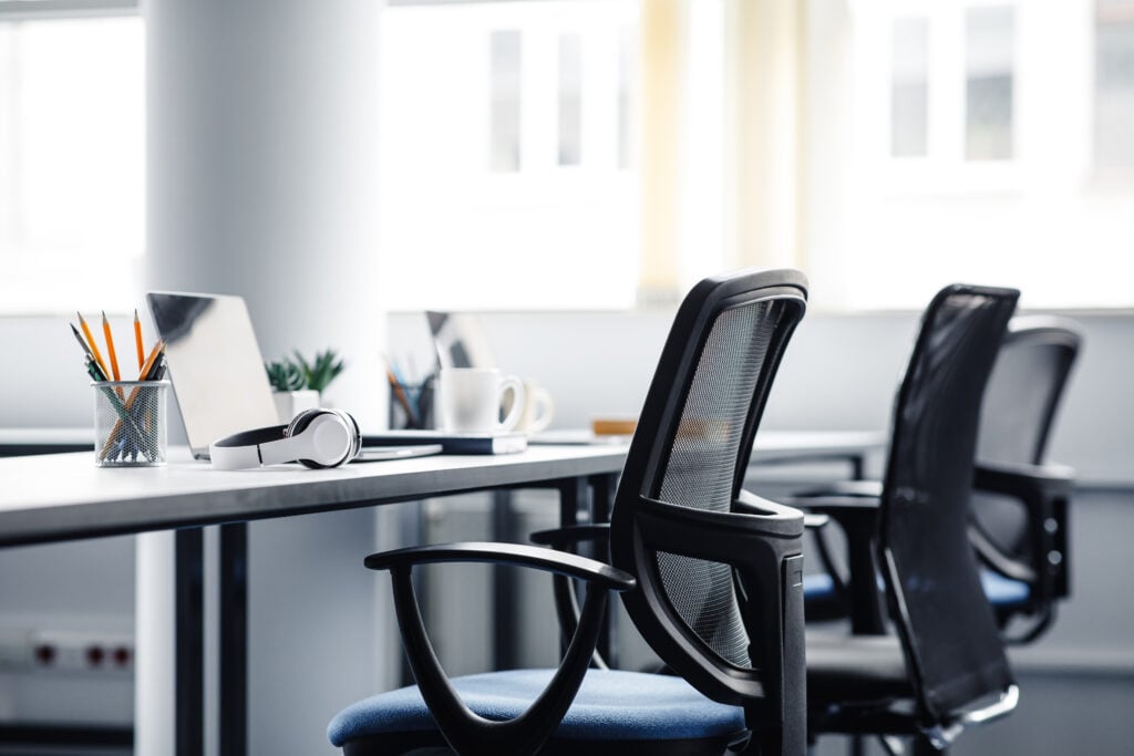 Modern office workspace with empty chairs ready for you to reseat, a laptop, headphones, a coffee mug, and a pencil holder on desks in a bright, well-lit room.