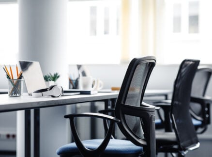 Modern office workspace with empty chairs ready for you to reseat, a laptop, headphones, a coffee mug, and a pencil holder on desks in a bright, well-lit room.