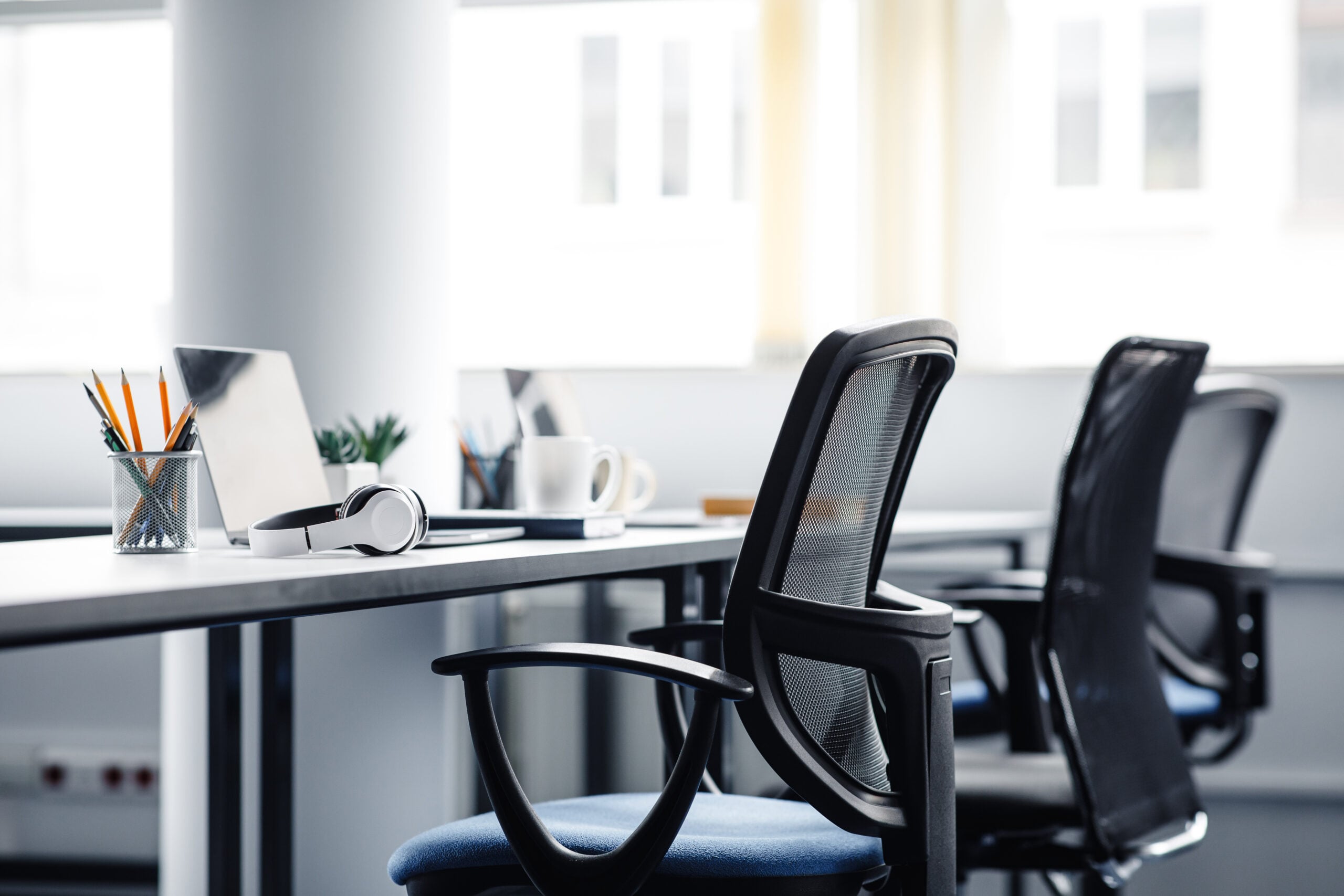 Modern office workspace with empty chairs ready for you to reseat, a laptop, headphones, a coffee mug, and a pencil holder on desks in a bright, well-lit room.