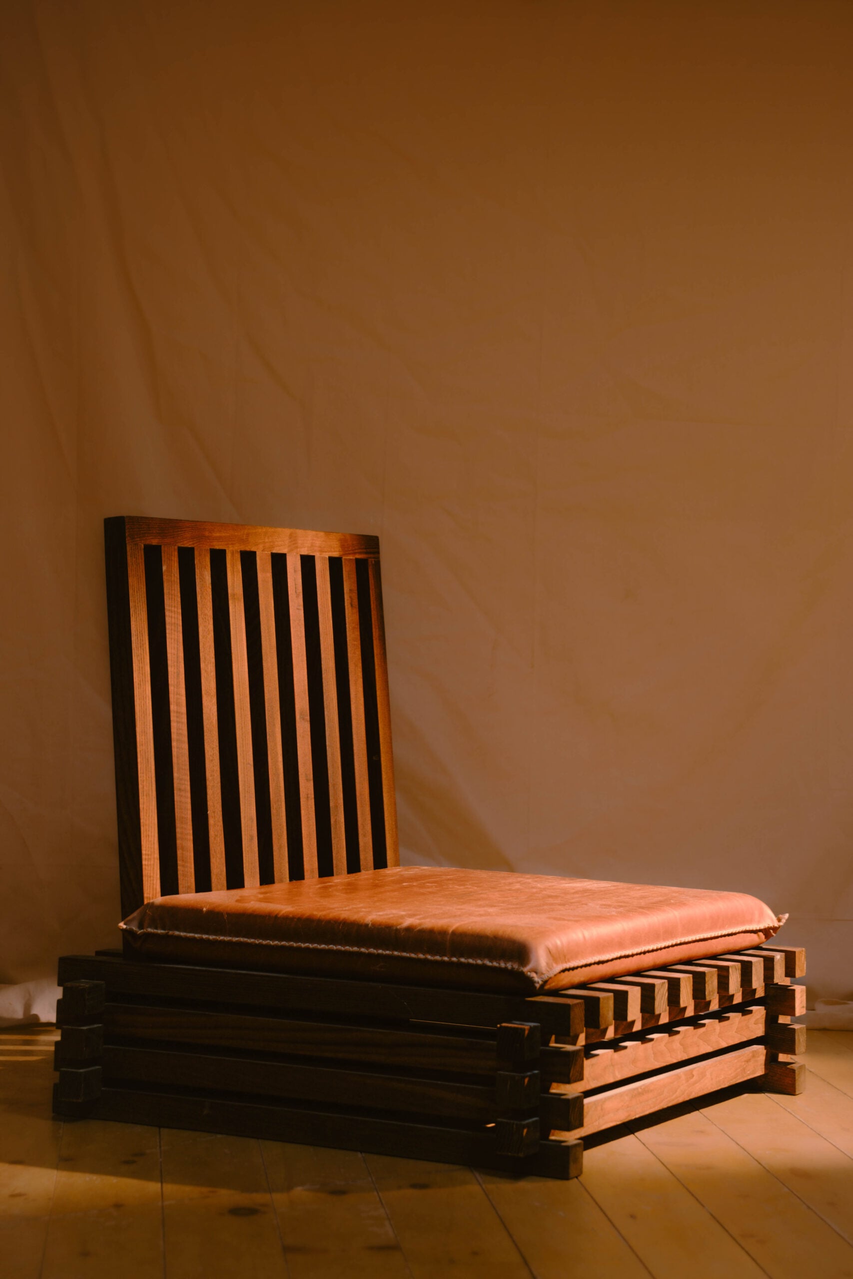 A low wooden chair with vertical slats on the backrest and a wide brown leather seat cushion, complemented by rich textiles, placed on a wooden floor against a beige backdrop.