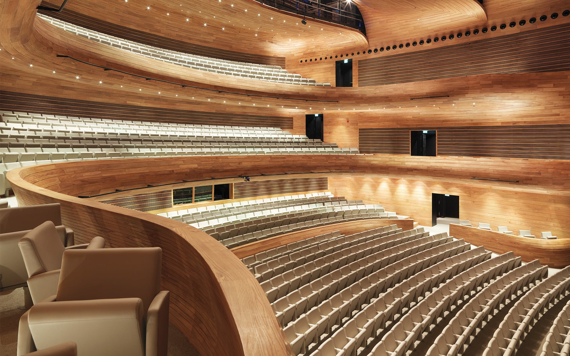 Modern concert hall interior featuring curved wooden walls, multiple balconies, and rows of light-colored seats, all empty—showcasing a stunning use of natural material in its elegant design.