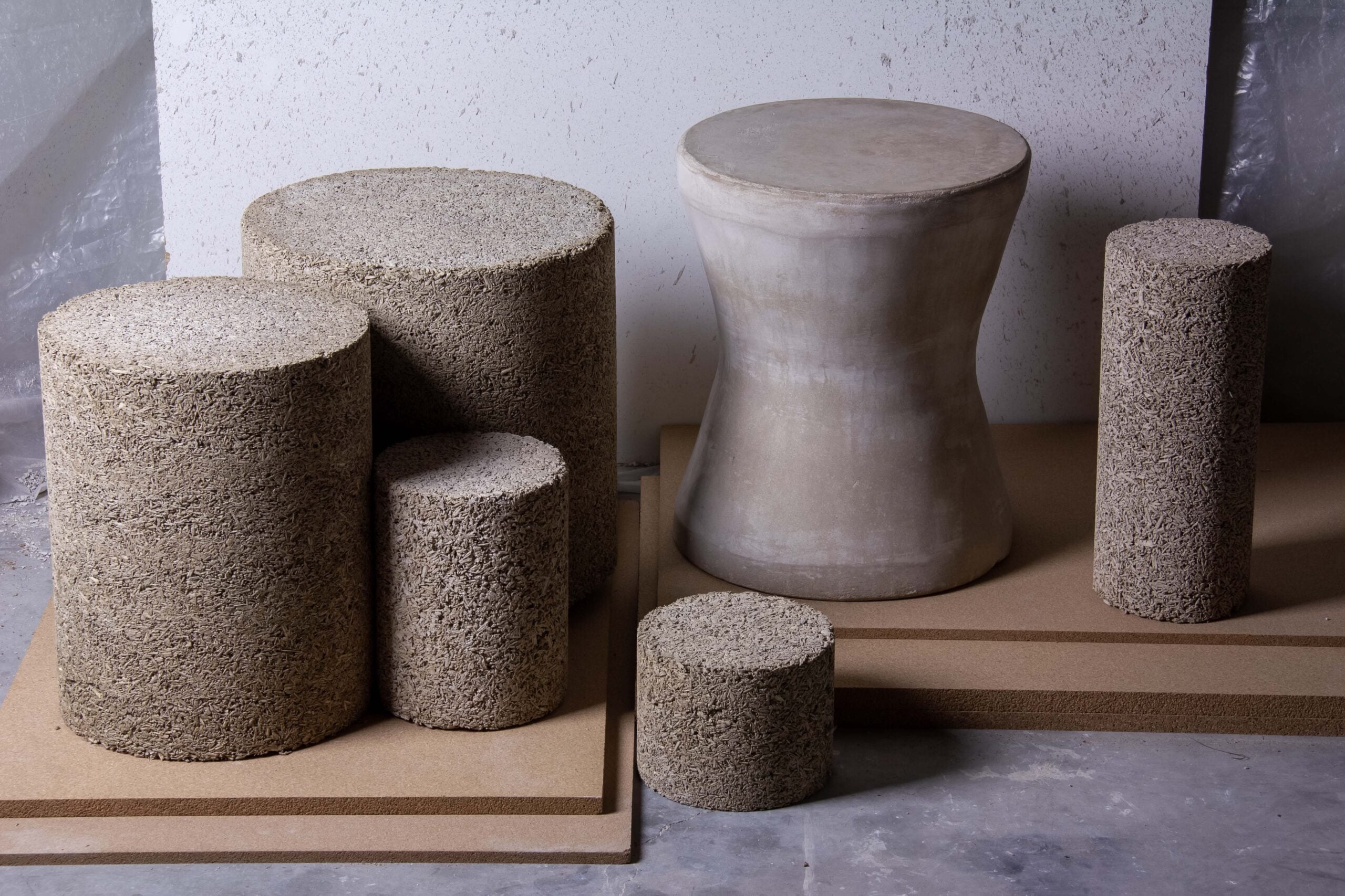Cylindrical and hourglass-shaped blocks, made of a rough, stone-like material, are arranged on rectangular boards against a textured wall, creating a striking contrast with nearby textiles.