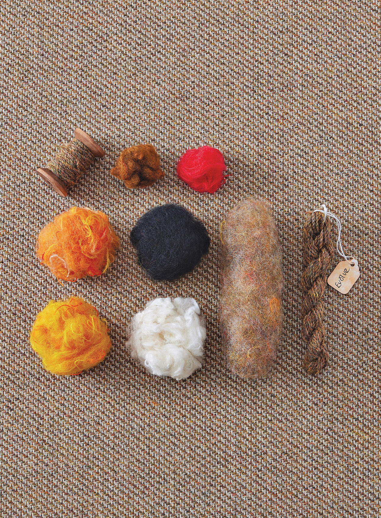 Various types and colors of wool yarn and fiber material, including spools, loose fibers, and a skein, arranged on a textured brown fabric background.