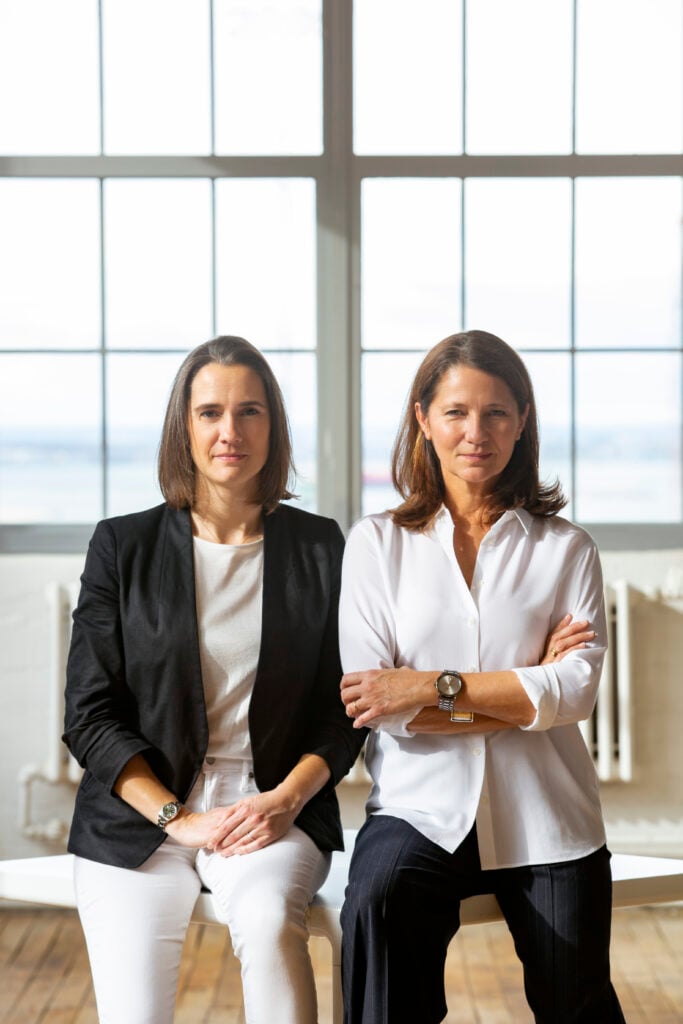 Two women sit side by side in front of large windows at ICFF, both dressed in business attire—one in a black blazer, the other in a white shirt—confidently looking directly at the camera.
