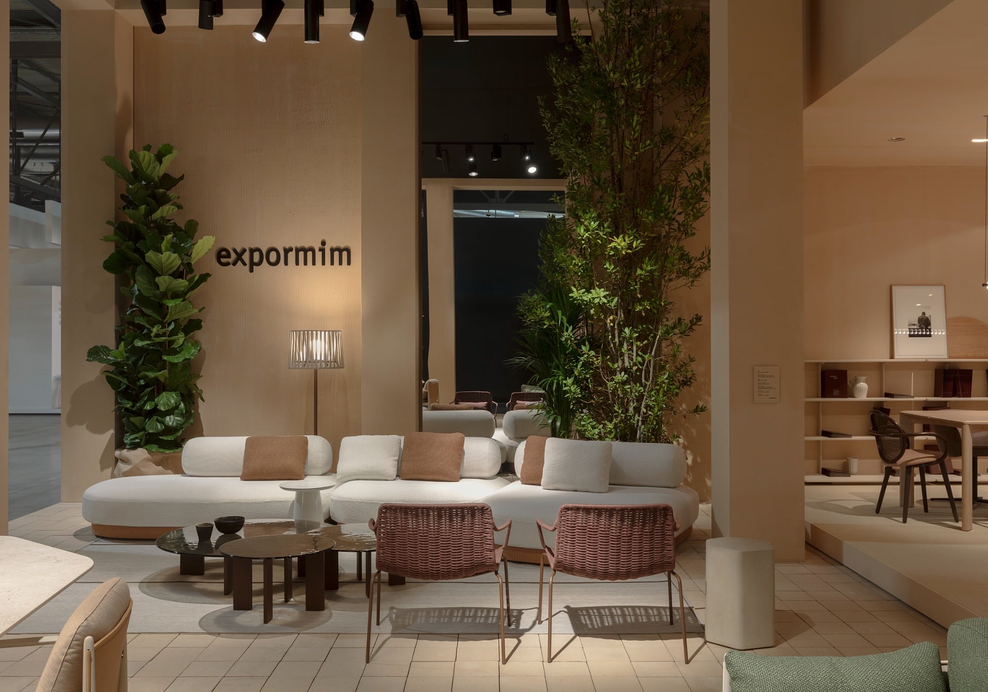 Modern living room showroom with a white sectional sofa, brown cushions, wicker chairs, green plants, and neutral-toned decor under soft lighting. Highlighting sustainable products inspired by Milan Design Week, "expormim" is displayed on the wall.