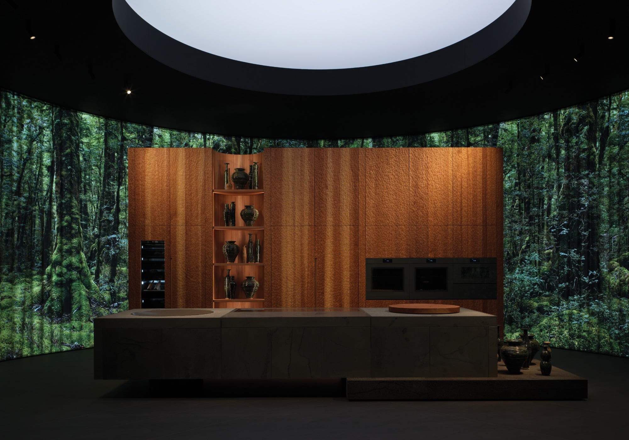 A modern kitchen with wood paneling and built-in appliances is set against a curved backdrop displaying a dense forest scene, inspired by sustainable products showcased at Milan Design Week, beneath a large circular skylight.
