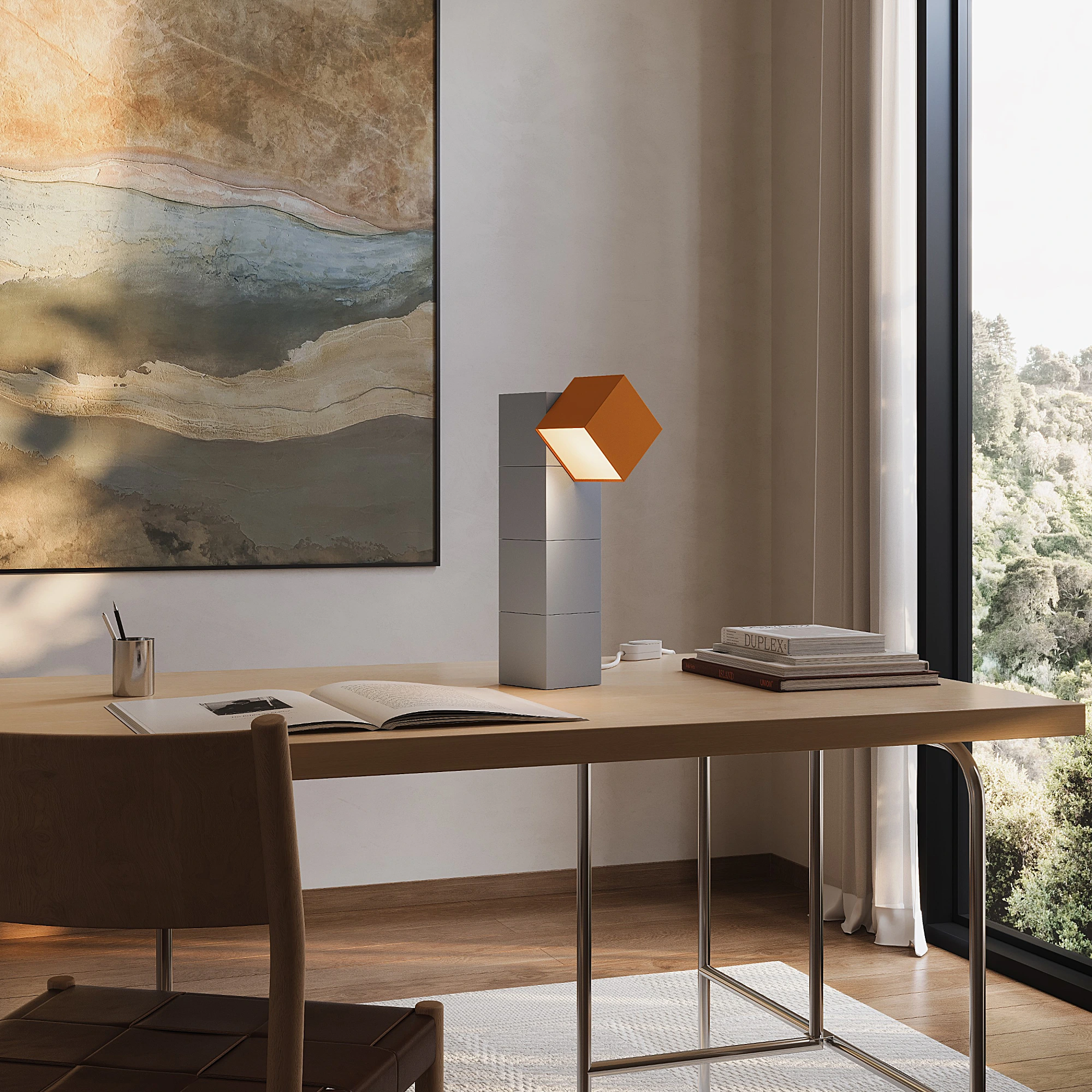 A modern desk with an open book, a geometric orange and gray lamp inspired by ICFF designs, a chair, large abstract wall art, and a window overlooking trees.
