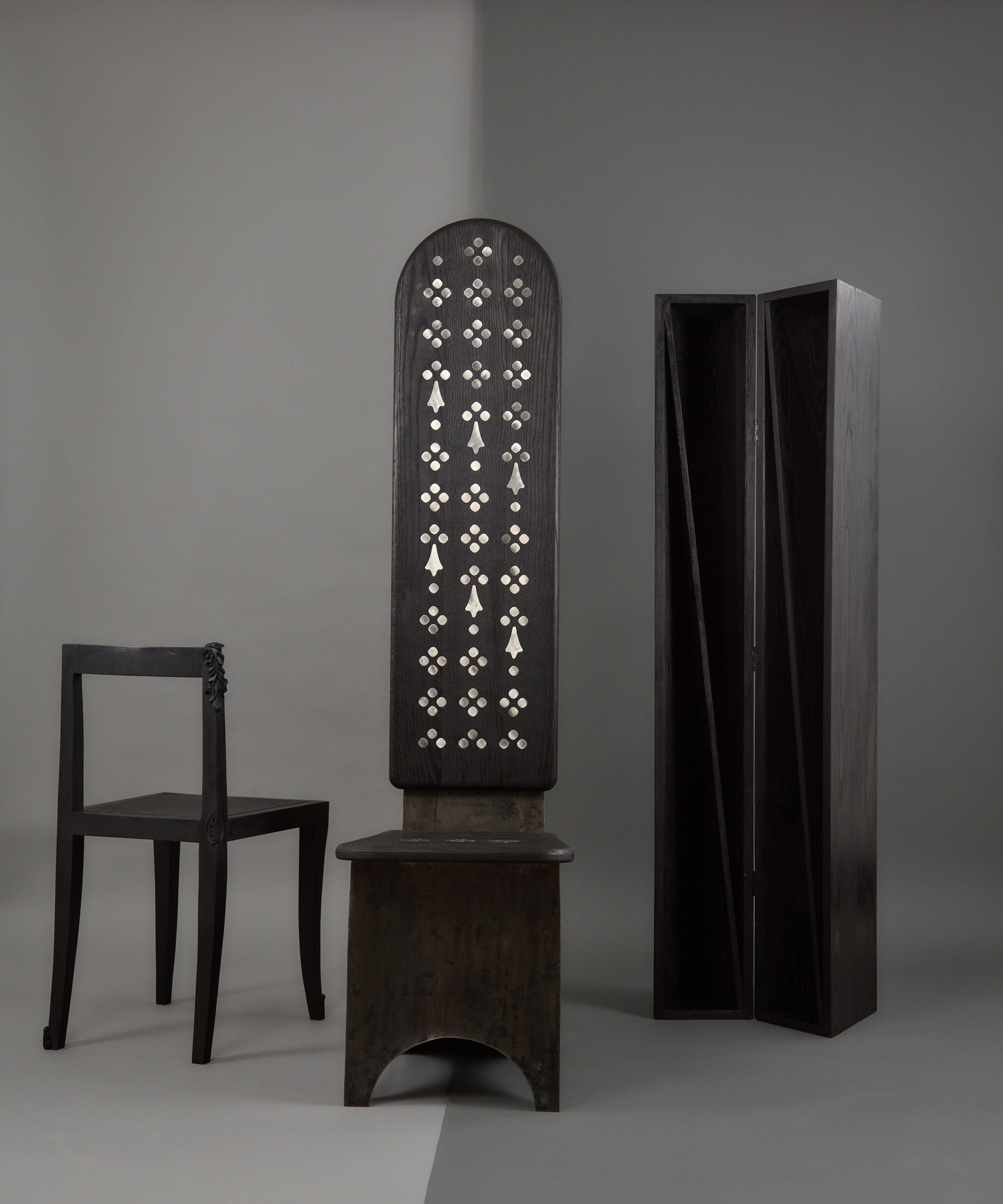 Three dark wooden furniture pieces—a simple chair, an arched-back chair with patterned inlay, and a tall, narrow folding screen—are set against a neutral background, complemented by subtle touches of textiles.