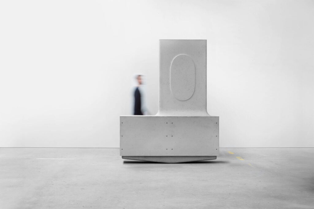 A person in motion walks past a large, abstract, gray concrete sculpture with geometric shapes—an embodiment of organic architecture—in a minimal, empty room.