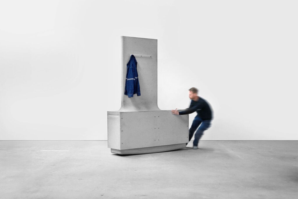 A person pushes a large, gray, L-shaped concrete structure—reminiscent of organic architecture—with a blue jacket hanging on a hook against a white wall.
