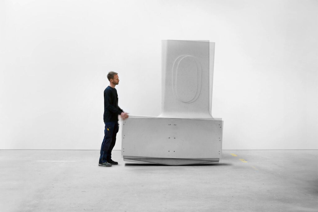 A man stands next to a large, white, geometric sculptural object in a minimal, empty room with a plain white wall, evoking the clean lines and harmony of organic architecture.