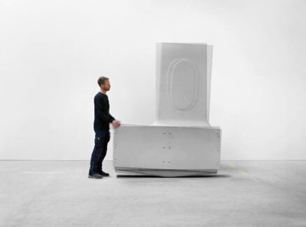 A man stands next to a large, white, geometric sculptural object in a minimal, empty room with a plain white wall, evoking the clean lines and harmony of organic architecture.