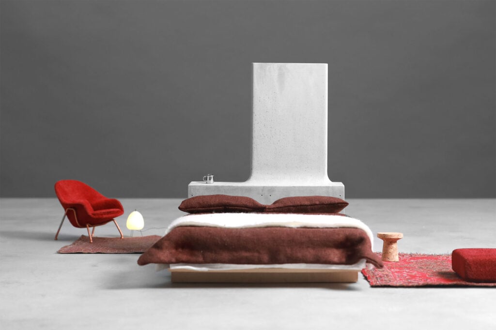 A minimalist bedroom scene featuring organic architecture with a bed, red armchair, small table, rug, and a white lamp, all set against a serene gray background.