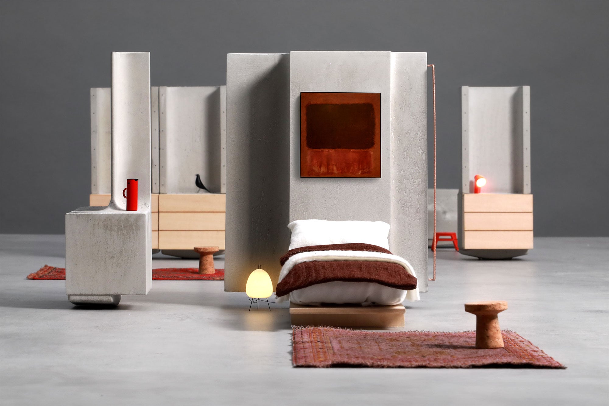 Minimalist modern bedroom setup featuring organic architecture elements with concrete walls, a single bed, rugs, small tables, lamps, and abstract artwork—all arranged on a gray floor with a neutral background.
