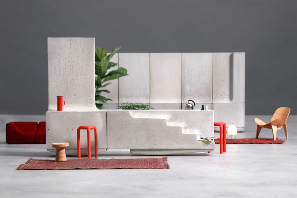 A modern, minimalist kitchen inspired by organic architecture, featuring concrete surfaces, red stools, a red rug, a wooden chair, and a plant in the background.