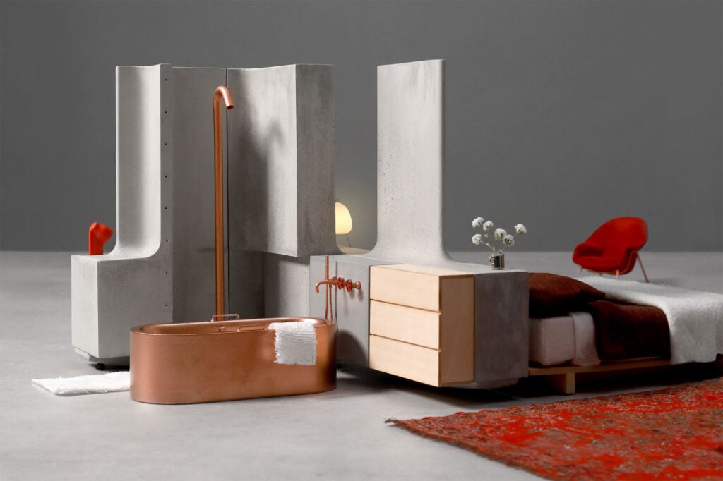 Modern minimalist bedroom and bathroom setup features organic architecture with concrete partitions, a copper bathtub, wooden bed and dresser, red chair, and red patterned rug on a gray floor.