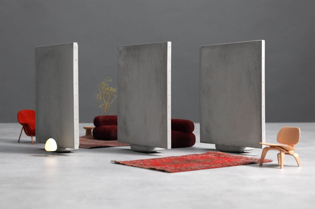 Three large, vertical concrete slabs evoke organic architecture as they stand on rugs, dividing a minimalist room with modern chairs, a small lamp, a plant, and a gray background.