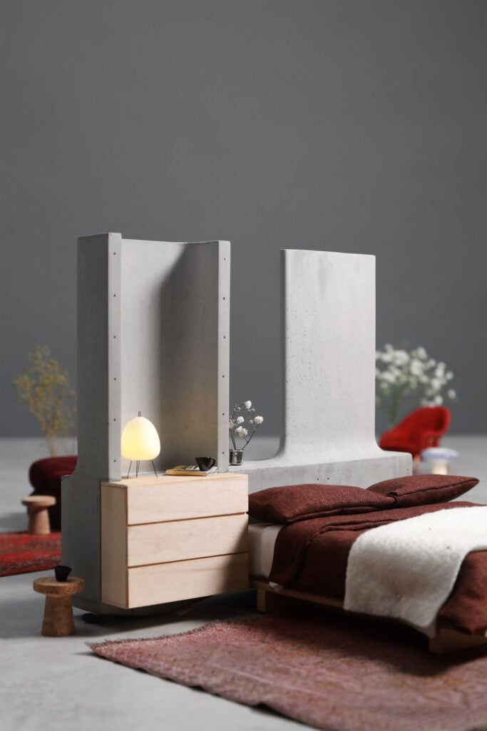 A modern bedroom with a minimalist design, featuring a concrete room divider and wooden nightstand, embraces organic architecture through natural materials. The bed with brown and white bedding stands out against a calming gray background.