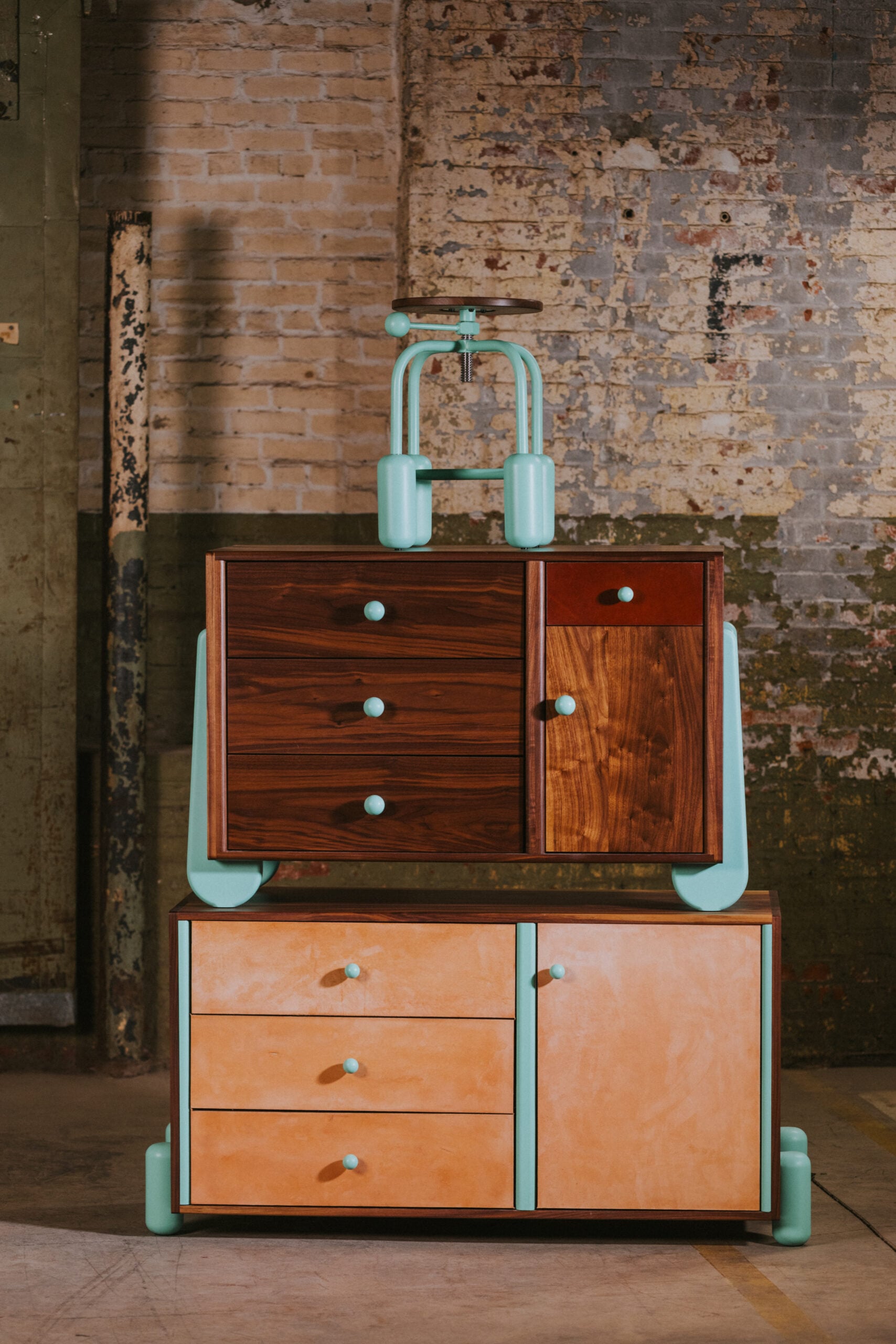 Two wooden cabinets, one stacked atop the other, feature blue handles and legs, set against an old brick and industrial wall background, complemented by artisanal textiles draped nearby.