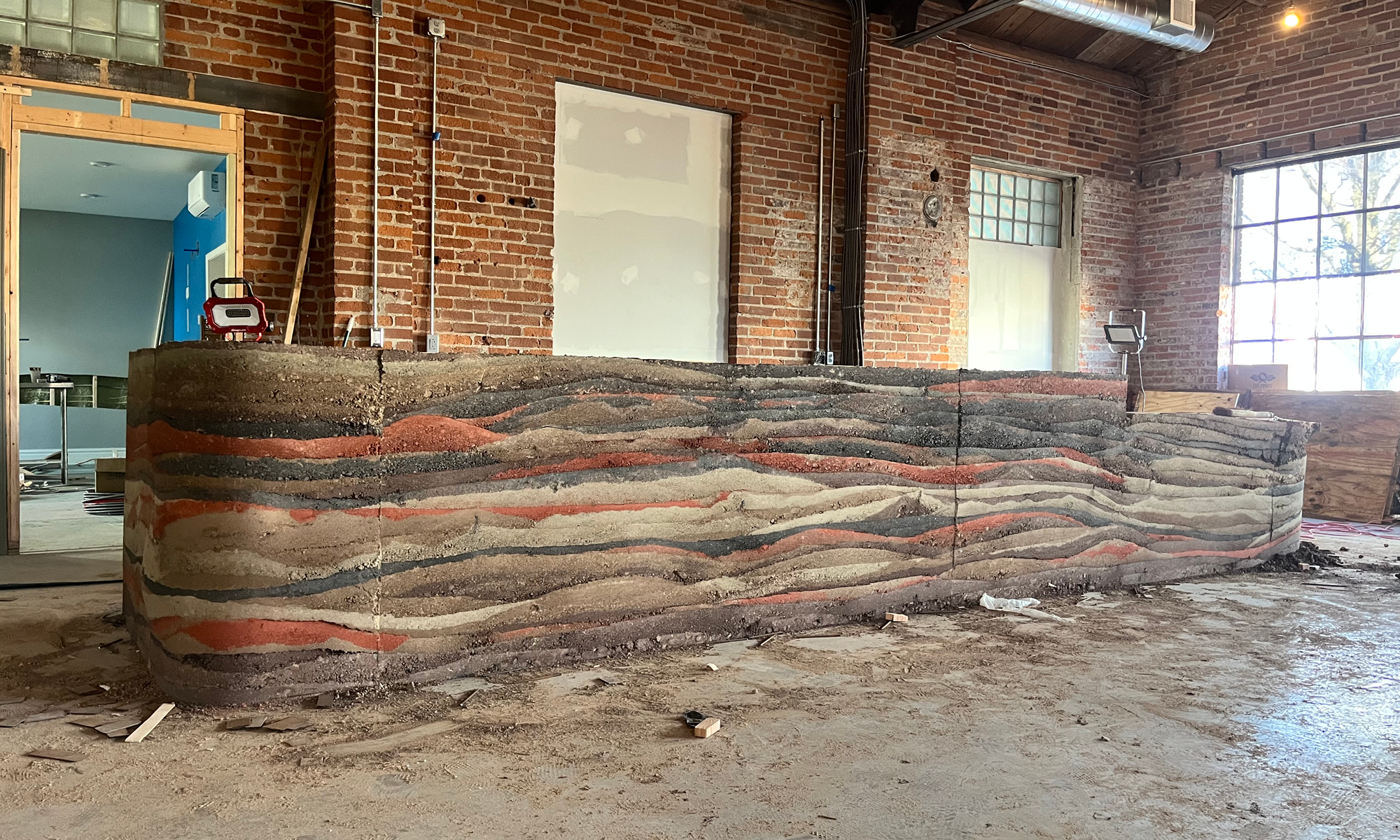 A curved rammed earth counter with layered earth tones stands in a partially constructed room, its design reflecting both natural beauty and the sustainability roadblocks faced during eco-friendly builds. Exposed brick walls and unfinished floors complete the setting.