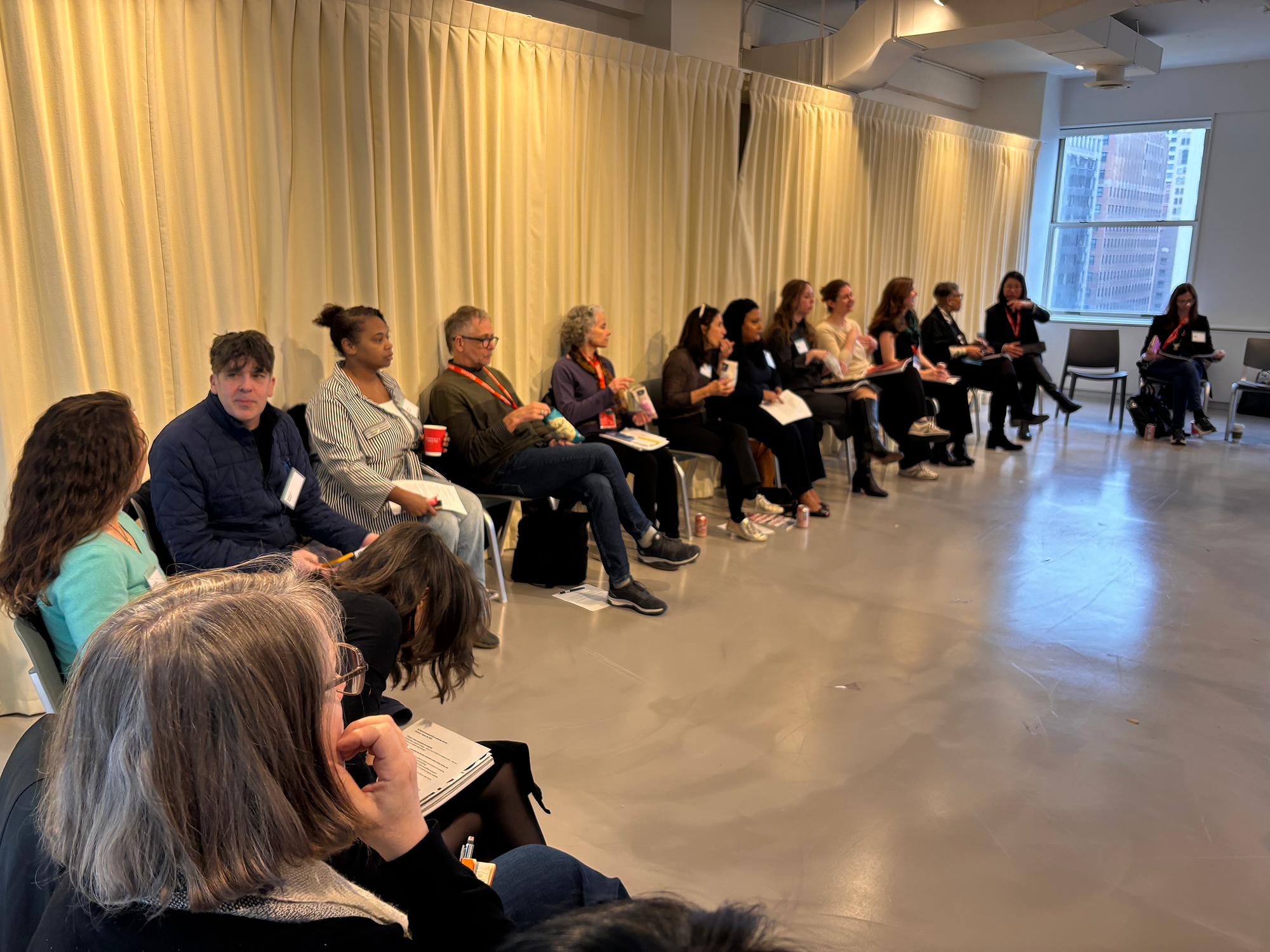 A group of people sit in a semicircle on chairs in a bright room with cream curtains, having a discussion or attending a workshop.