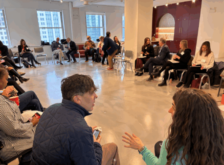 People sitting in a large circle in a brightly lit room, engaged in small group conversations; city buildings visible through the windows in the background.