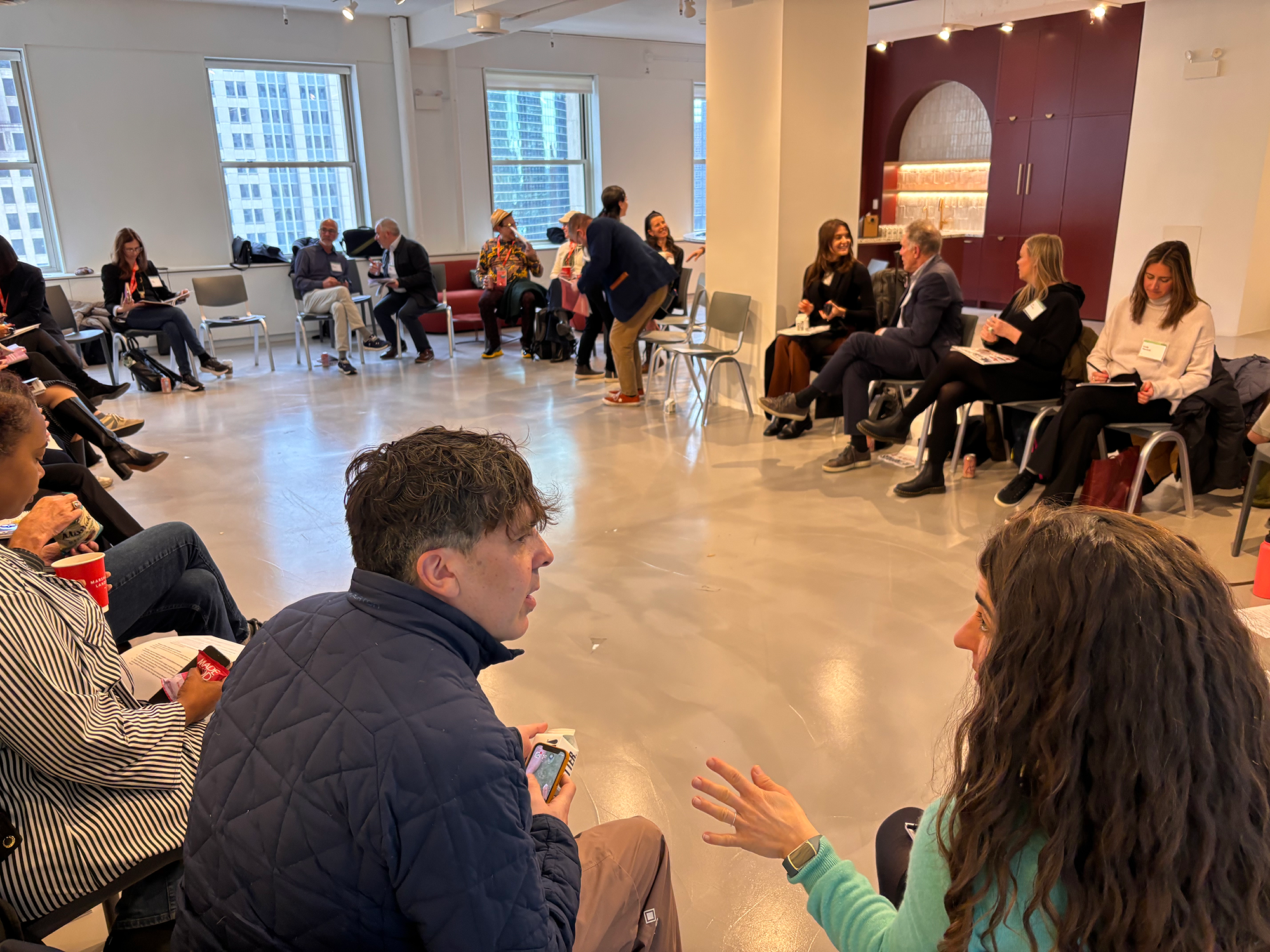 People sitting in a large circle in a brightly lit room, engaged in small group conversations; city buildings visible through the windows in the background.