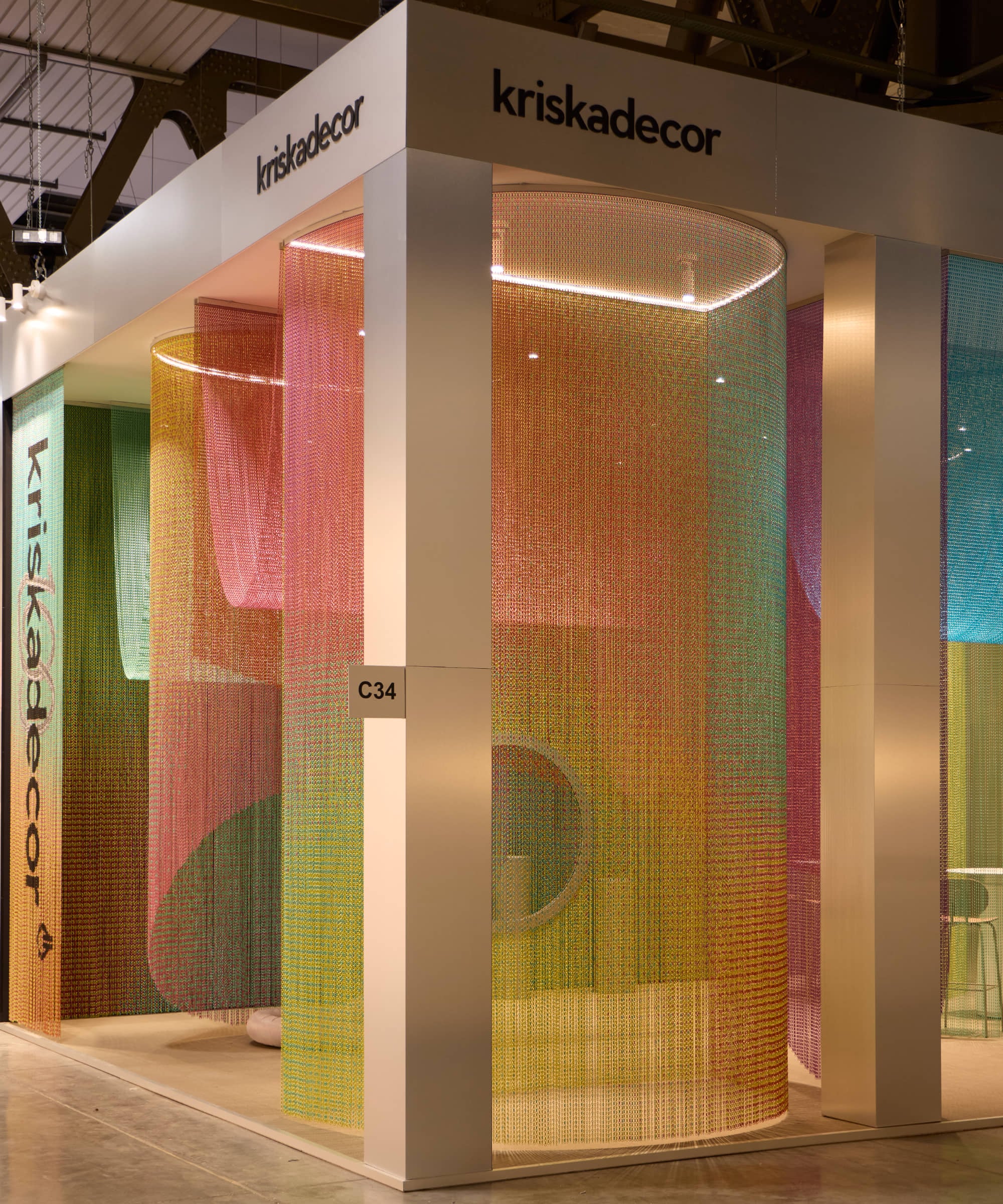 Exhibition booth with white framework and multicolored metal chain curtains, labeled "kriskadecor," showcases sustainable products at Milan Design Week, with a sign marked C34 and visible seating inside.