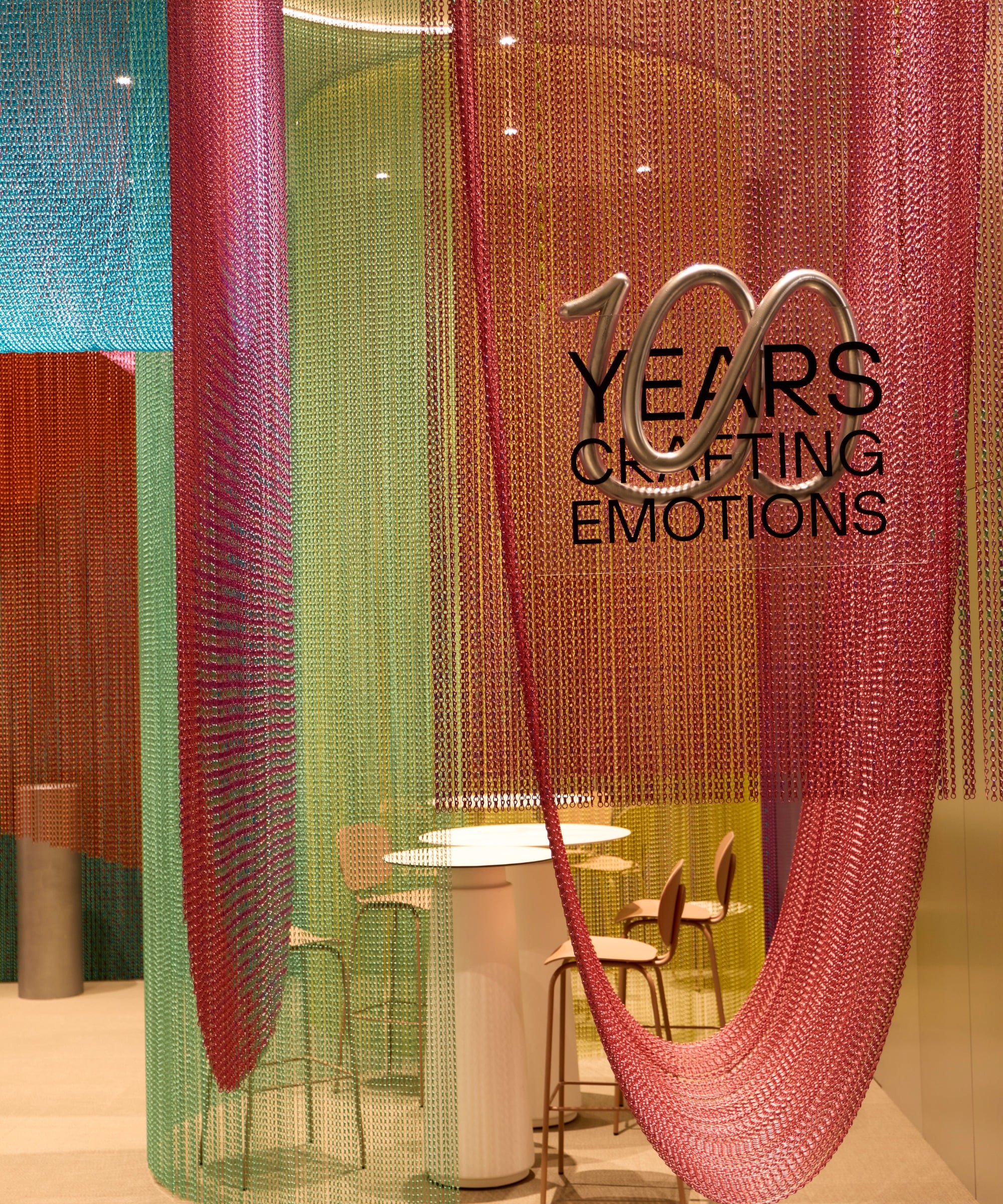 A modern seating area with colorful hanging chains, round tables, chairs, and a wall sign that reads "100 Years Crafting Emotions," inspired by Milan Design Week's focus on sustainable products.