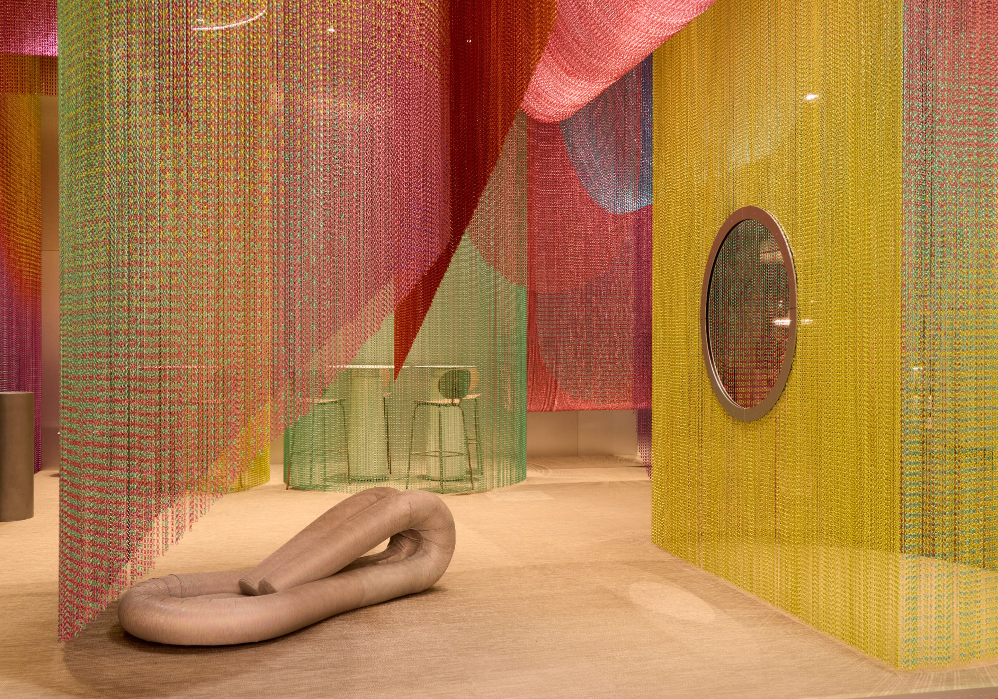 Colorful chain curtains in yellow, pink, and green hang from the ceiling, with a beige sculptural seat and a round window—creating a modern interior inspired by Milan Design Week and sustainable products.