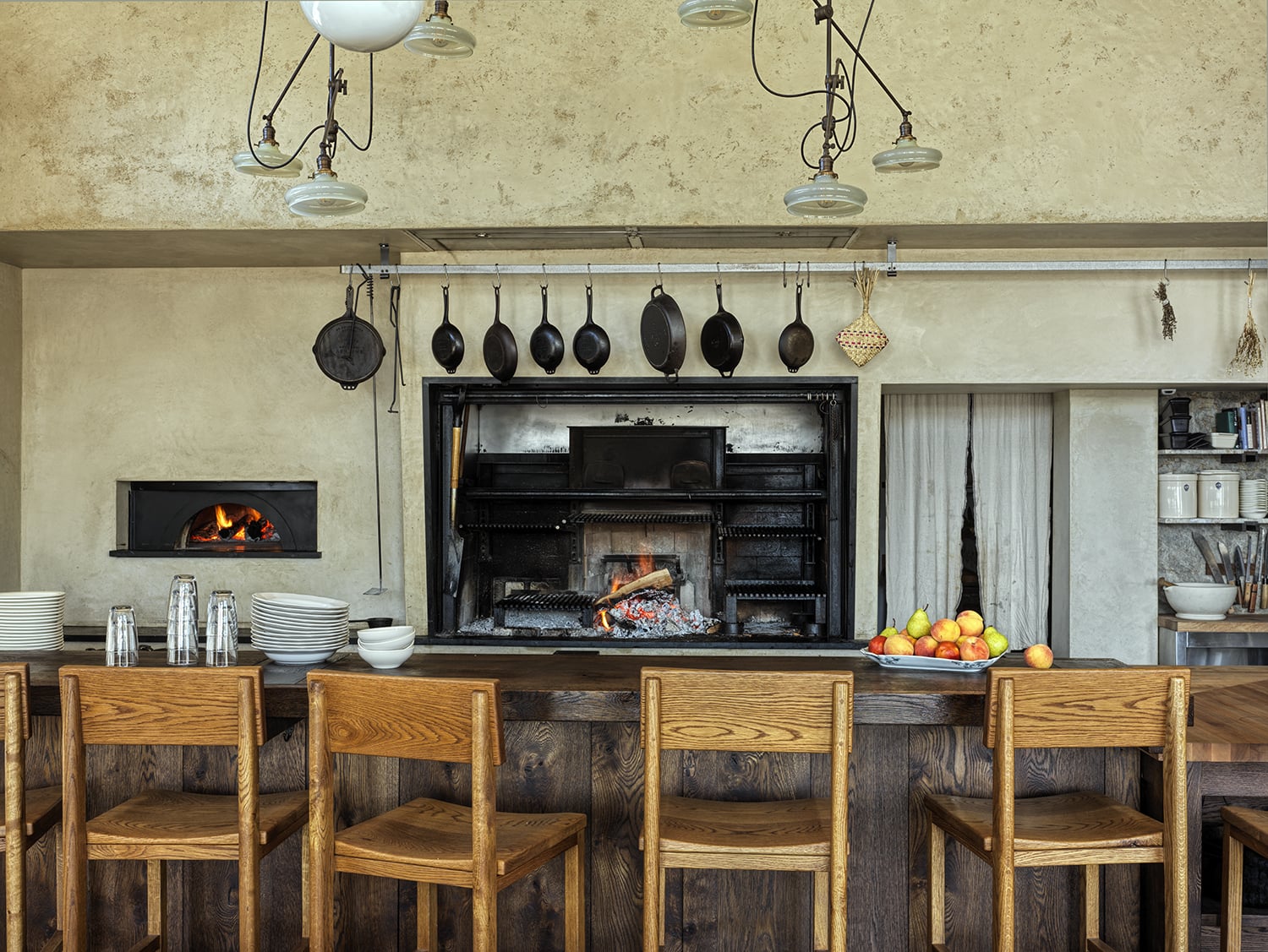 A rustic kitchen where lovers unite, featuring a wood-fired oven, open hearth grill, hanging cookware, wooden chairs at the counter, and a bowl of apples on the countertop.