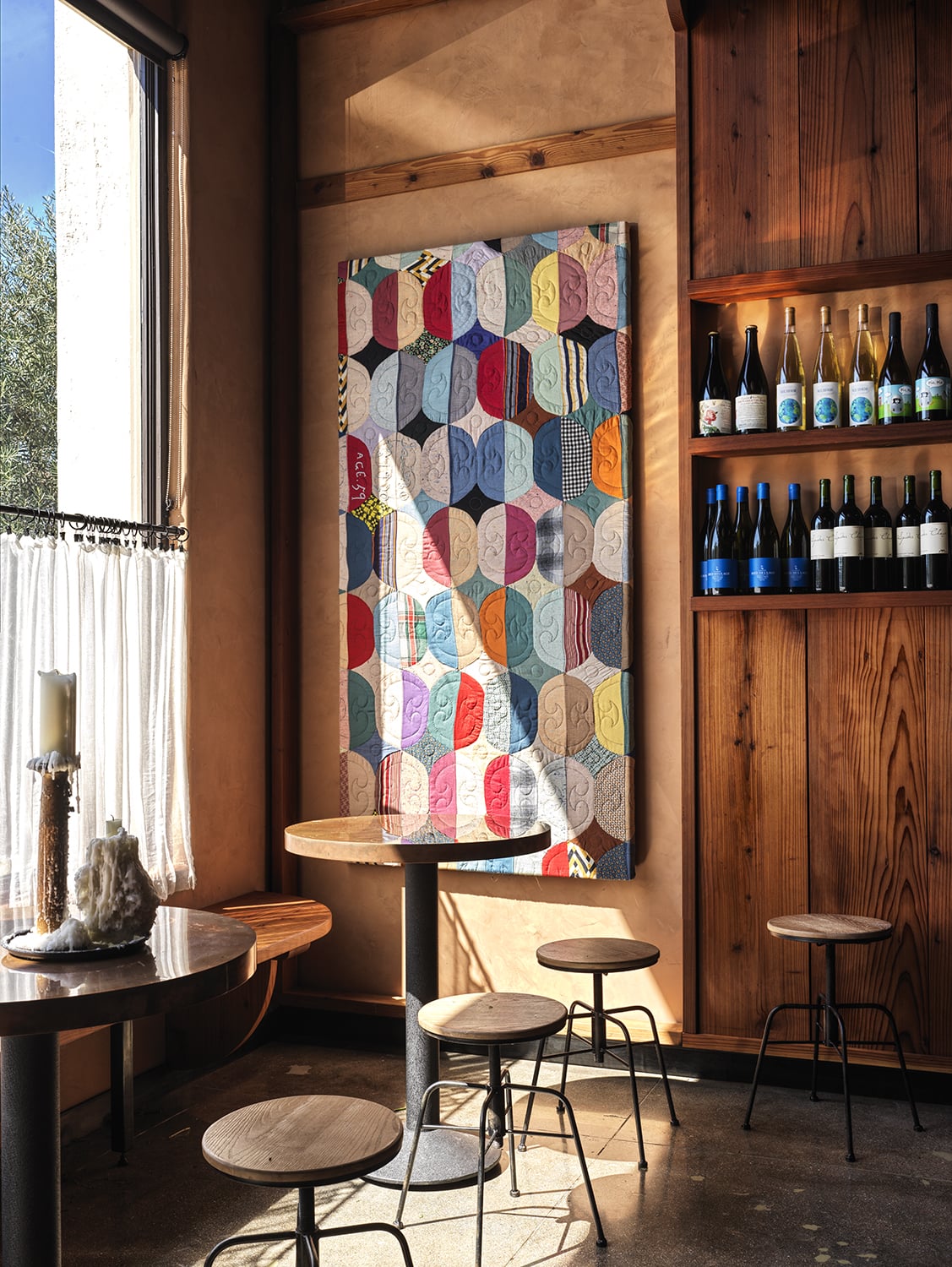 A cozy cafe corner with round wooden tables, stools, a wall-mounted wine shelf, and a colorful geometric-patterned quilt hanging on the wall—an inviting spot where lovers unite over warm drinks and good conversation.