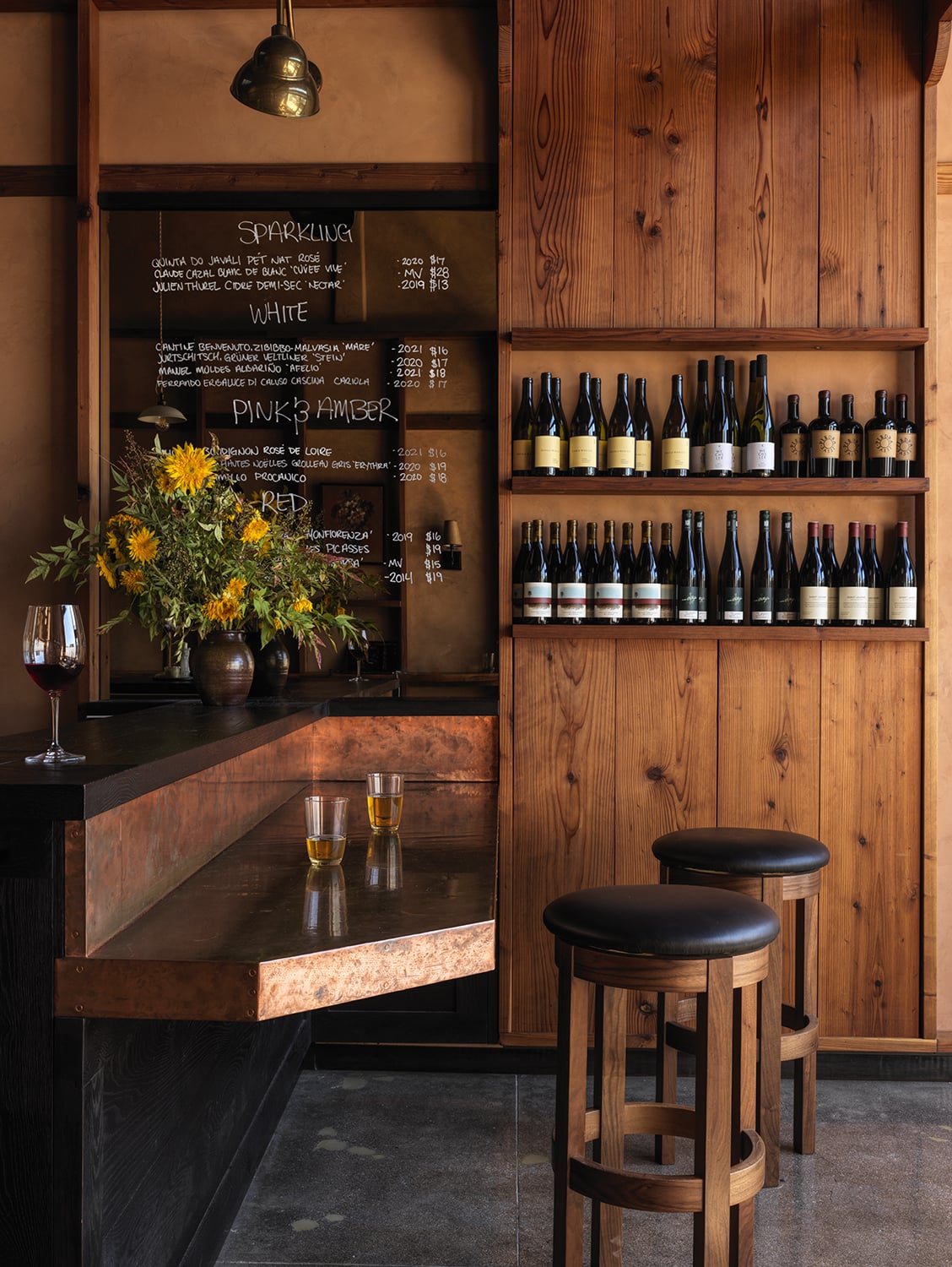 A cozy wine bar with wooden walls, two stools, a counter set for lovers unite over glasses of white wine, a bouquet of flowers, shelves of wine bottles, and a chalkboard menu.
