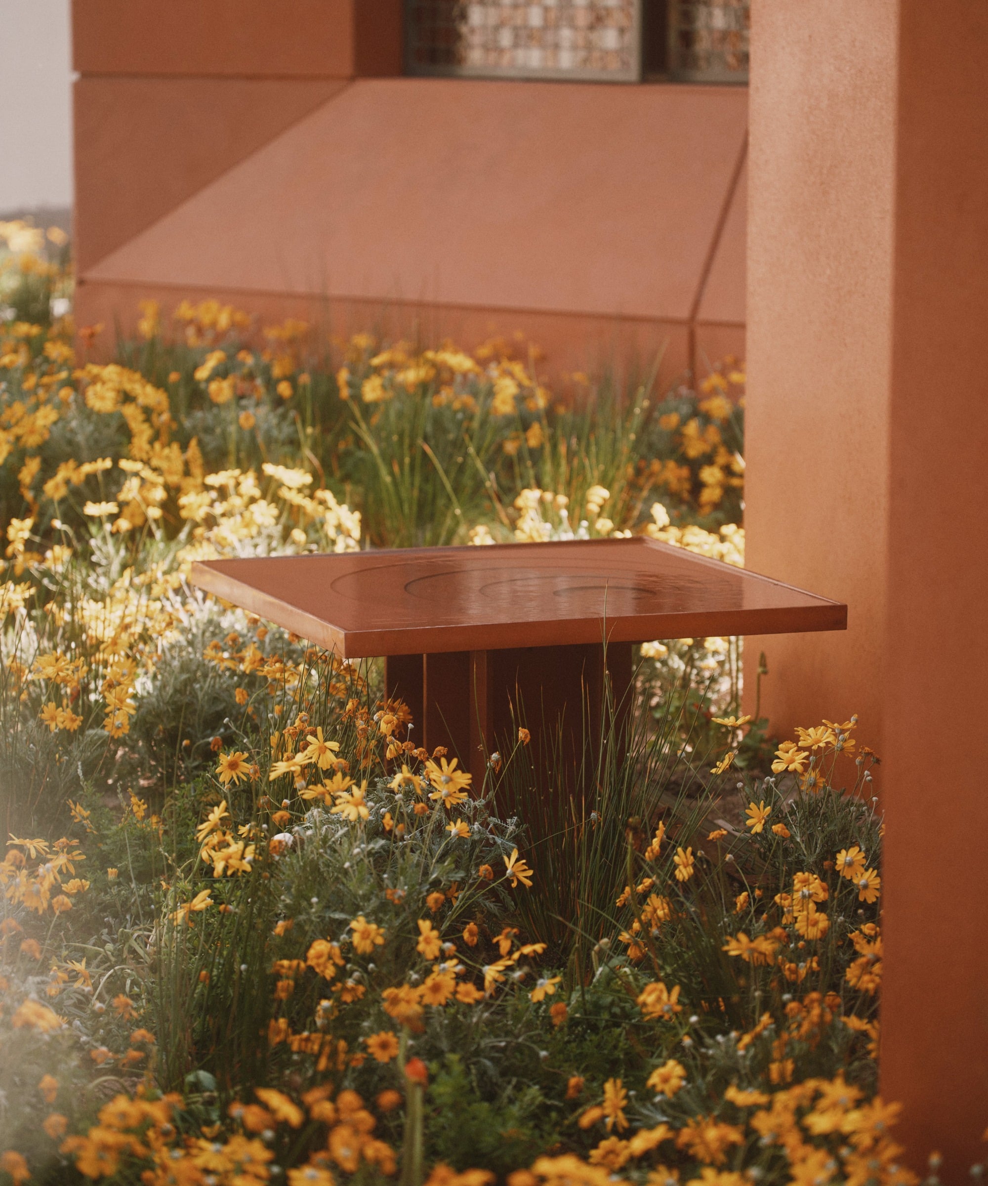 A square, rust-colored metal table—echoing the spirit of Milan Design Week sustainable products—stands among tall green grasses and yellow wildflowers next to an orange building.