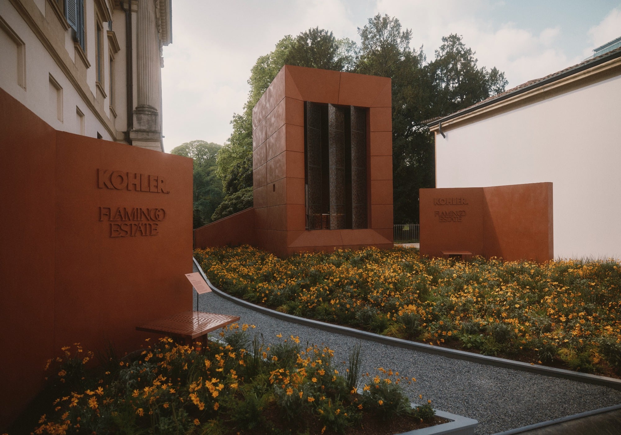 A modern rust-colored pavilion sits among yellow flowers and gravel paths at Milan Design Week, with “KOHLER” and “FLAMINGO ESTATE” signage highlighting their sustainable products on two nearby walls.