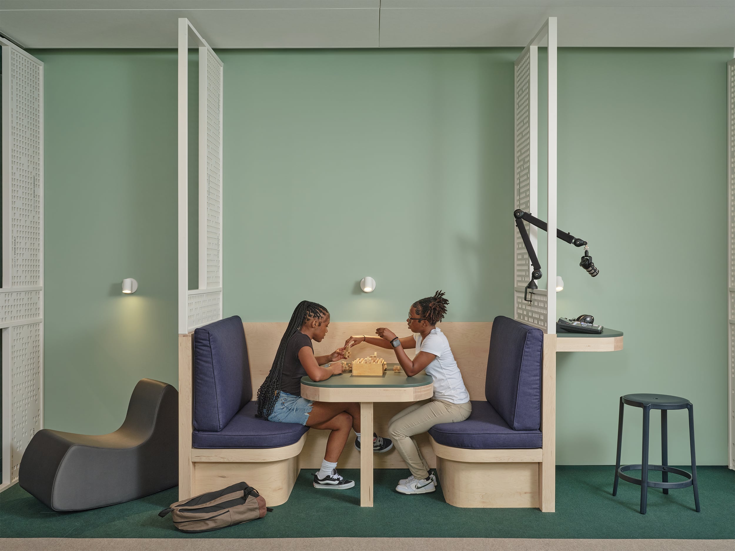 Two people sit across from each other at a booth-style table playing chess in a modern room with green walls and minimalist furniture.