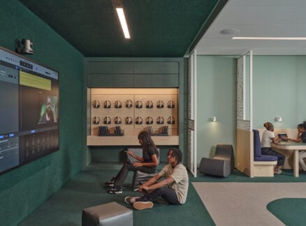 People in a modern study space use digital devices; two sit on the floor by a large screen, another sits nearby, and two more work at a round table. Headphones and tablets are stored on shelves.